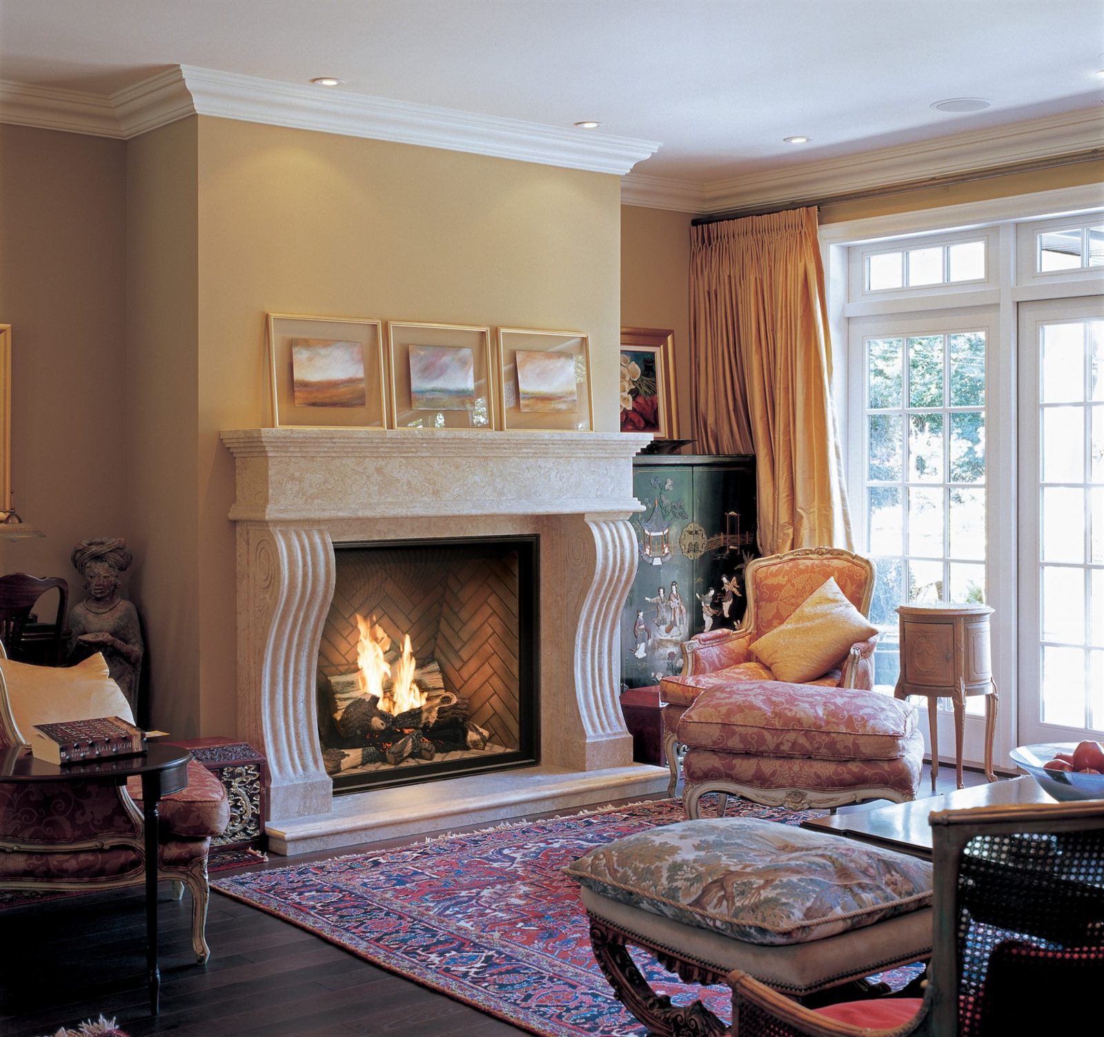 Town & Country traditional gas fireplace with a split-log fire on a herringbone brick liner, set into a carved cream stone mantel in a formal sitting room with damask armchairs, a Persian rug and tall French windows