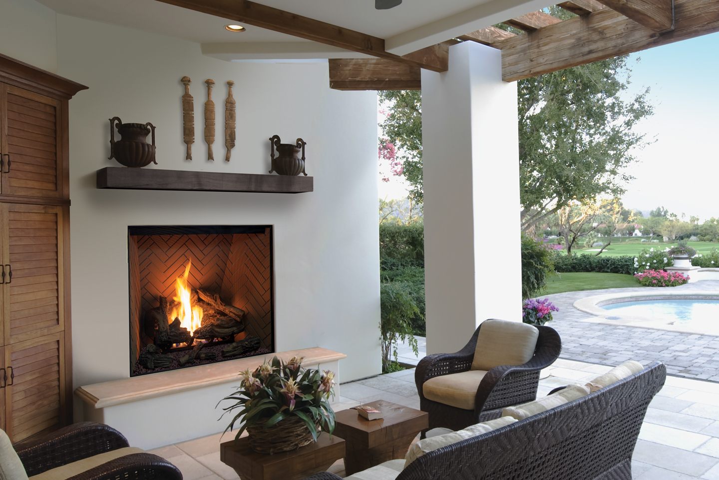 Urbana U44 Outdoor gas fireplace with a traditional log fire on a herringbone brick liner, set into a white wall beneath a timber mantel on a covered patio with wicker armchairs, a pergola and a pool and lawn beyond