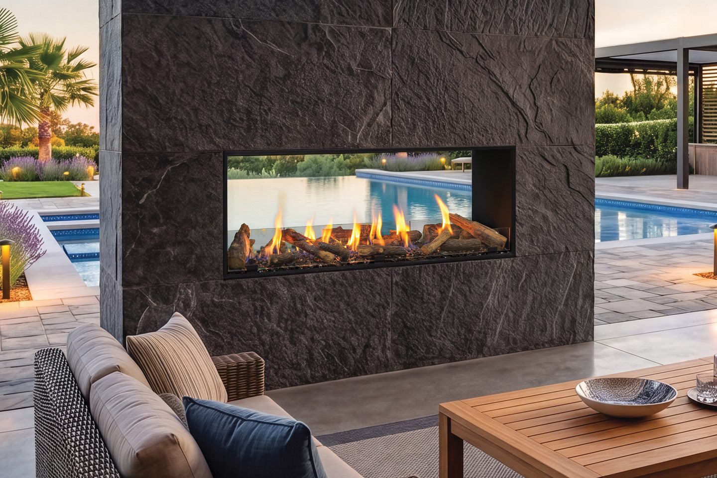 Urbana U60 Outdoor Linear gas fireplace set into a dark split-stone pier beside a swimming pool at dusk, the fire seen straight through to the water and lavender beyond, a wicker sofa and teak table in front