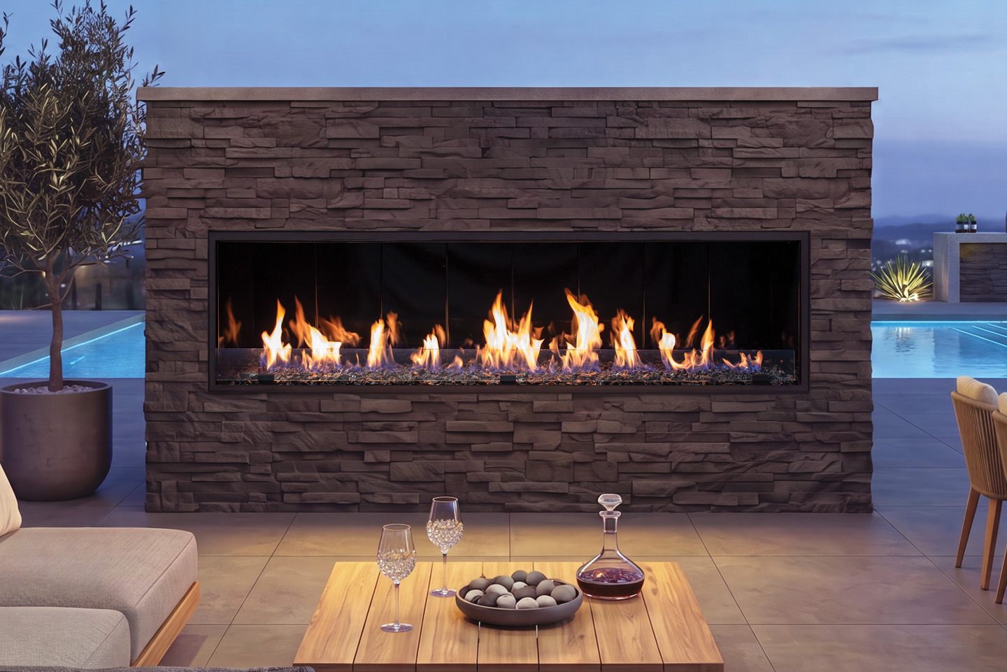 Urbana U74 Outdoor Linear gas fireplace, a long ribbon of flame over crushed glass set into a brown stacked-stone wall between a potted olive tree and a rooftop pool at blue hour, with a teak table and wine glasses in front