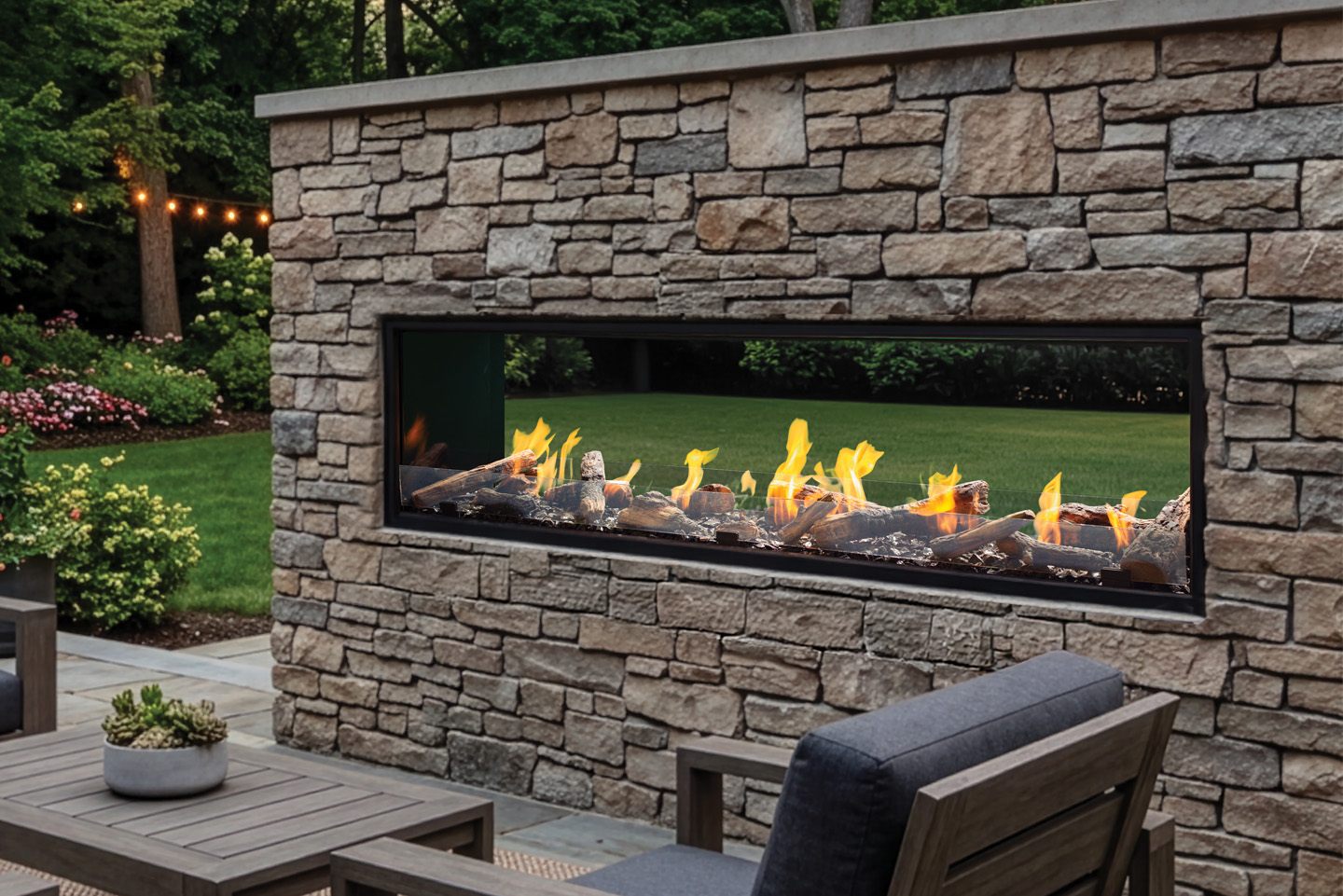 Urbana U74 Outdoor Linear gas fireplace installed straight through a dry-stacked stone garden wall, driftwood logs burning behind a low glass guard with a lawn, flower beds and string lights visible through the fire on the far side