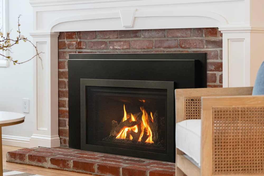 Valor G2 gas insert with a black front in a red brick fireplace beneath a white mantel with a panelled surround, a rattan armchair at the right