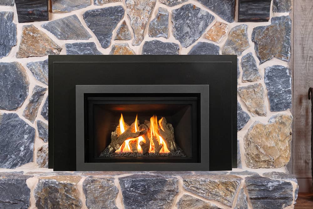 Valor G2 gas insert with a black three-sided front set into a rough grey and tan stone fireplace, a log fire burning behind the glass