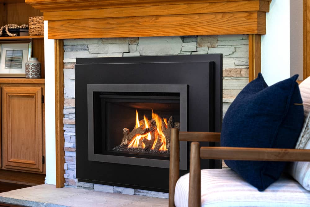 Valor G2 gas insert with a black four-sided front in a stacked-stone fireplace beneath an oak mantel, a navy cushion and a wooden armchair in the foreground