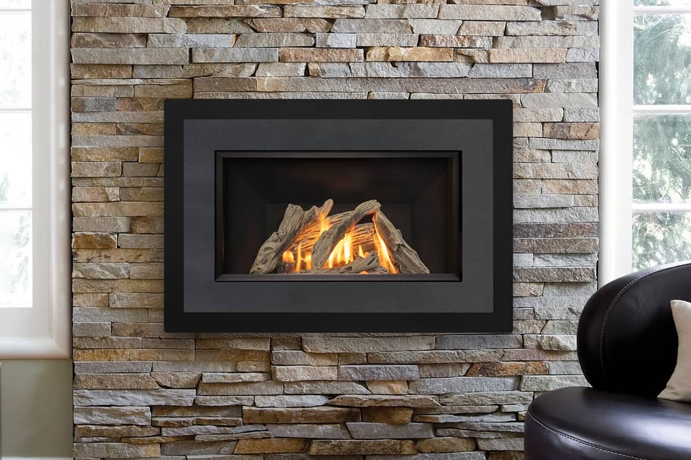 Valor Horizon with a pewter H4 Landscape front and a driftwood log set, set into a wall of stacked stone between tall windows beside a black leather chair