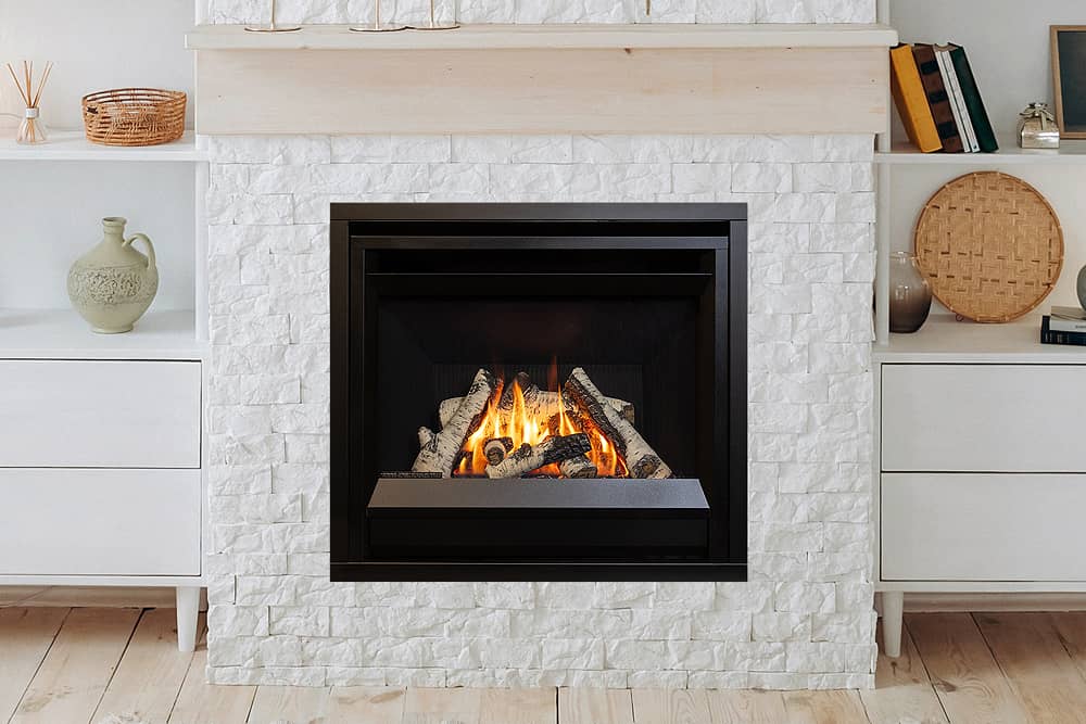 Valor Horizon gas fireplace with a birch log set and a plain black front, set into a wall of white stacked stone beneath a pale wood mantel between two white cabinets