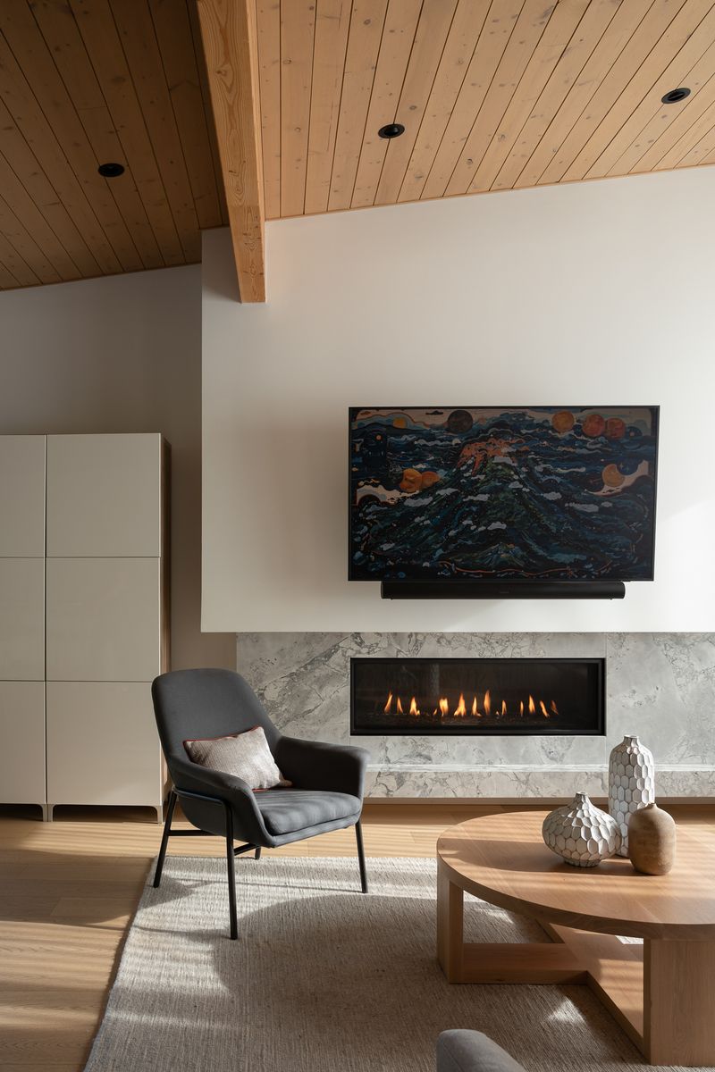 Valor L2 linear gas fireplace set low into a wide grey marble surround beneath a television, in a calm living room with a plank ceiling, a charcoal armchair and a round oak coffee table