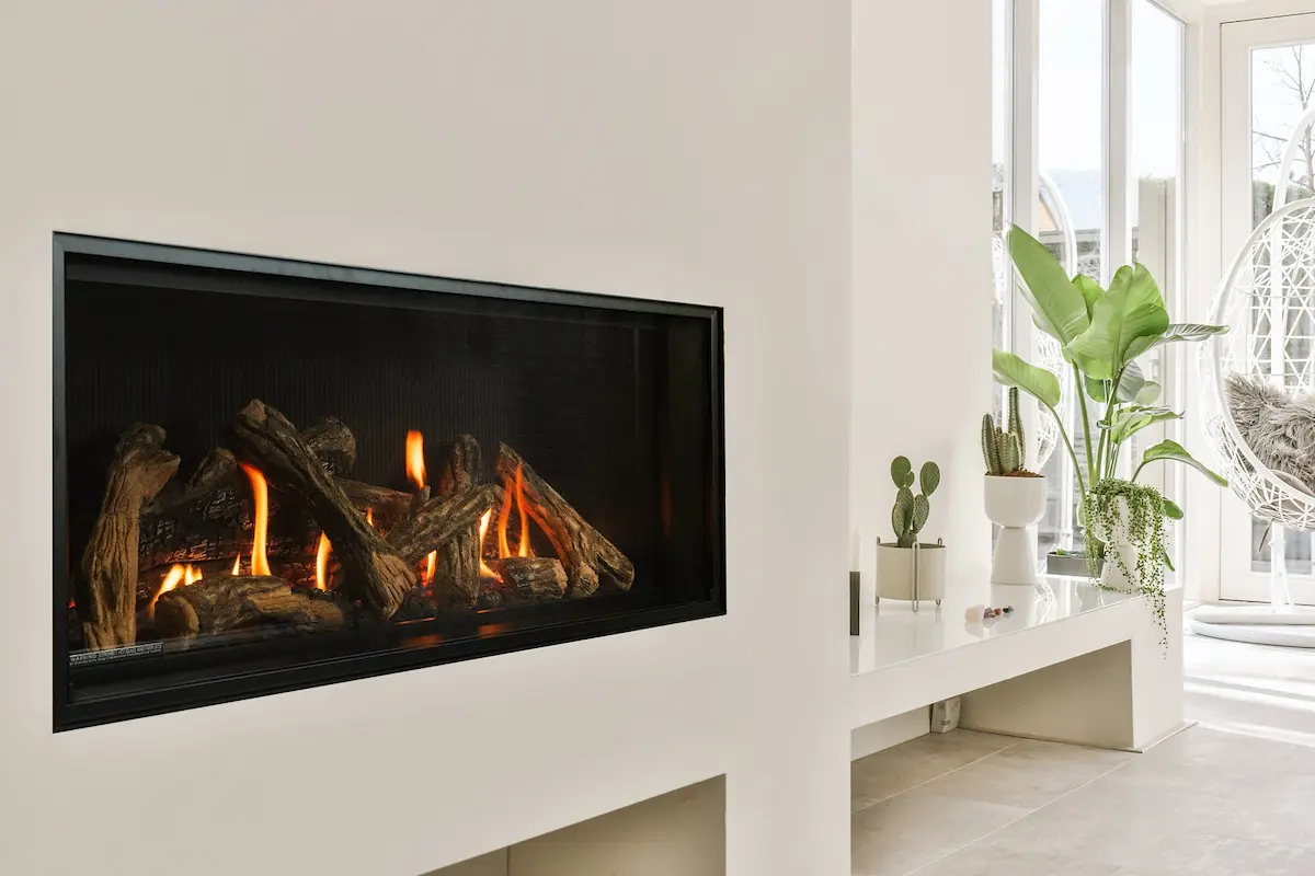 Valor LT2 Landscape gas fireplace set into a smooth white wall in a bright, glass-walled living room, a tall stack of logs burning behind edge-to-edge glass beside a long white bench with potted plants