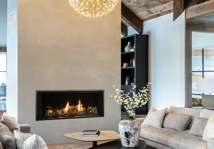 Valor LT2 Landscape gas fireplace with a split-log fire set into a tall plaster chimney breast, a low oak table and a pale linen sofa in front of it beneath a glass globe chandelier and reclaimed timber beams