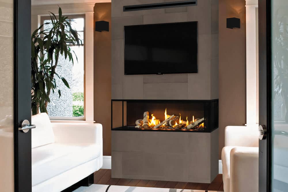 Valor LX2 three-sided gas fireplace with a birch log fire set into a grey stone pier between two rooms, glass on the front and both ends, a television and the slim HeatShift vent above, white sofas on either side