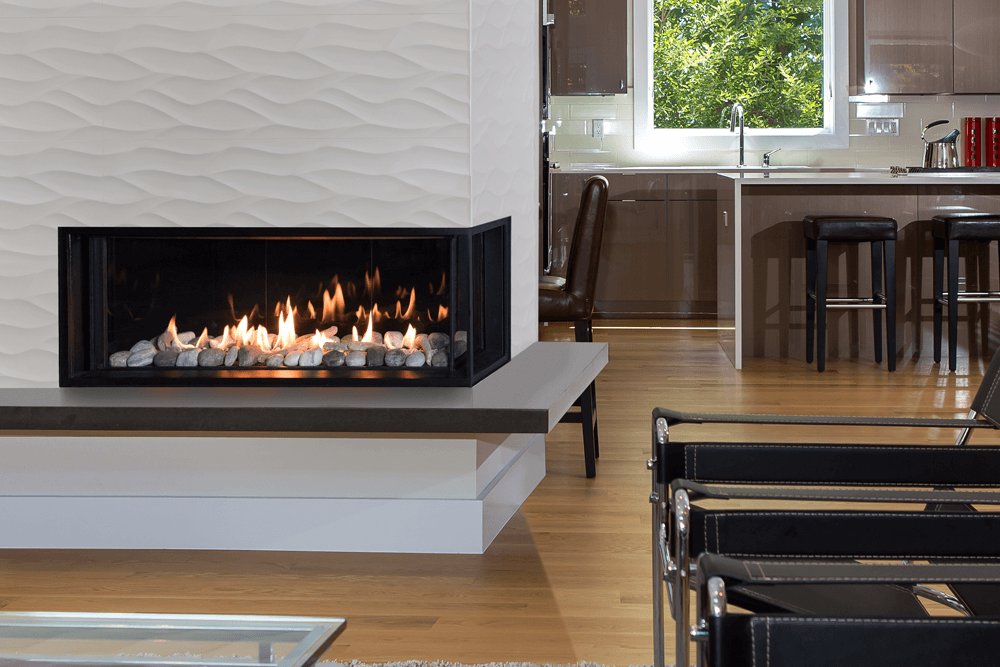 Valor LX2 corner gas fireplace with a rock and shale bed set into a rippled white tile wall at the end of a grey stone bench, glass on the front and the right-hand end, an open kitchen with bar stools beyond