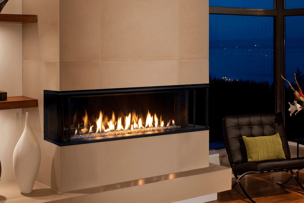 Valor LX2 gas fireplace with a Murano glass bed and reflective liner wrapping the corner of a pale stone pier at dusk, a long ribbon of flame beside a black leather chair and a window over a harbour